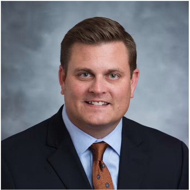 A professional headshot of a smiling man in a dark suit, light blue shirt, and a patterned brown tie against a gray studio…