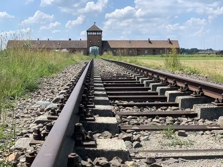 A low-angle shot of weathered train tracks leading toward the iconic brick gatehouse of Auschwitz-Birkenau concentration…