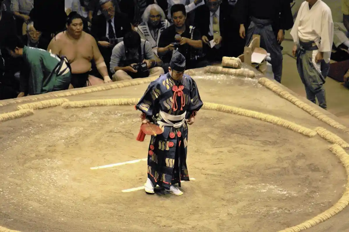 A sumo wrestler in traditional ceremonial attire bows deeply in the center of a sumo ring, surrounded by other wrestlers and…