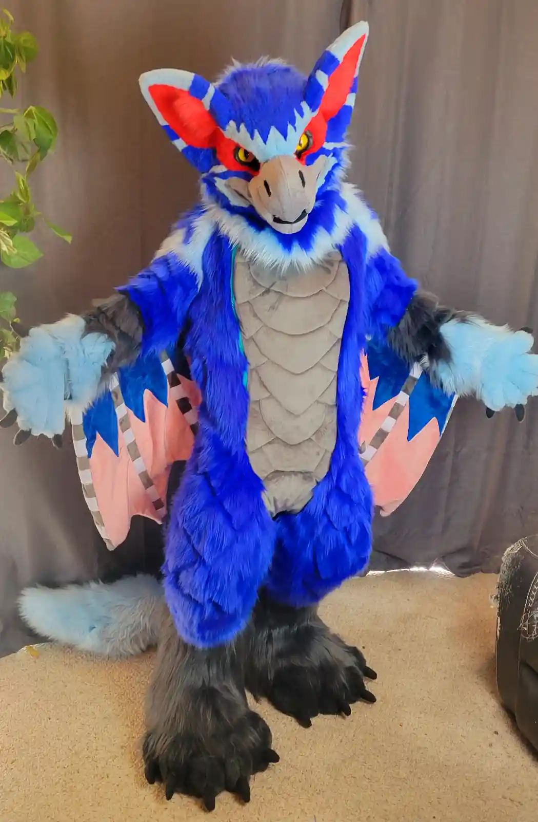 A person in a full-body blue, red, and white fursuit resembling a dragon or draconic creature stands with arms outstretched…