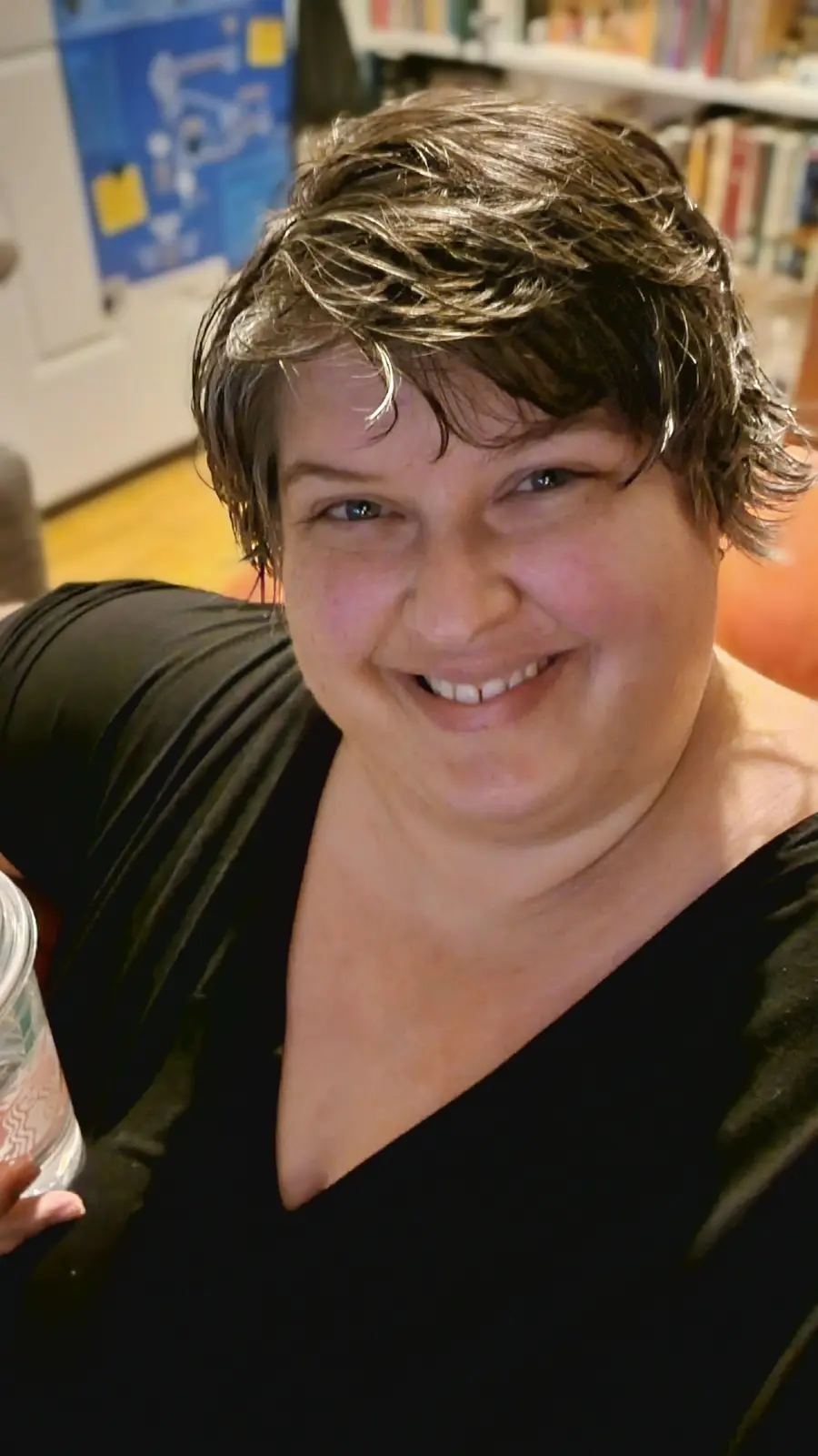 A person with short brown hair and a black top smiles while holding a glass, standing in front of a bookshelf and a blue…