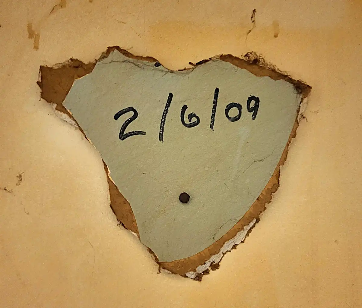 A close-up of a roughly heart-shaped hole punched through a beige wall, revealing a gray inner layer. The date '2/6/09' is…