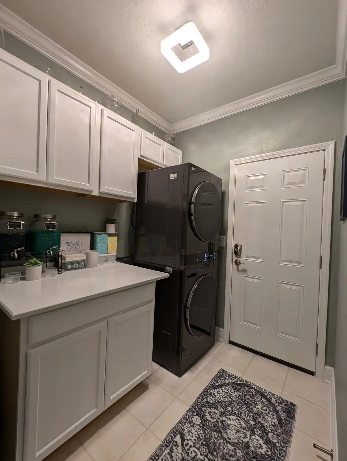 A clean, modern laundry room with white cabinets, a white countertop, and a stacked black LG washer and dryer unit. A white…