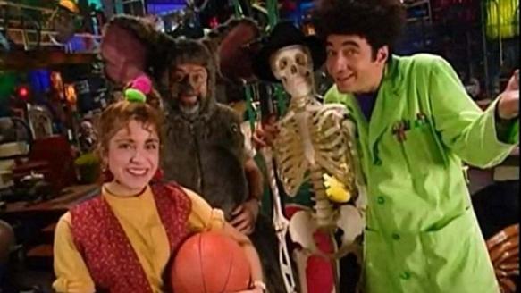 A group of people in a colorful, cluttered room, including a woman holding a basketball, a man in a green lab coat holding a…