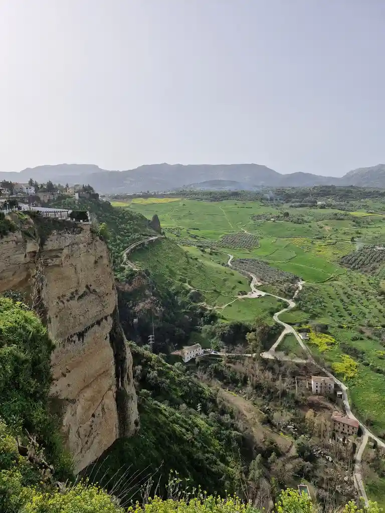 A scenic photo of a steep cliff overlooking a valley with winding roads, green fields, and distant mountains. The image has…