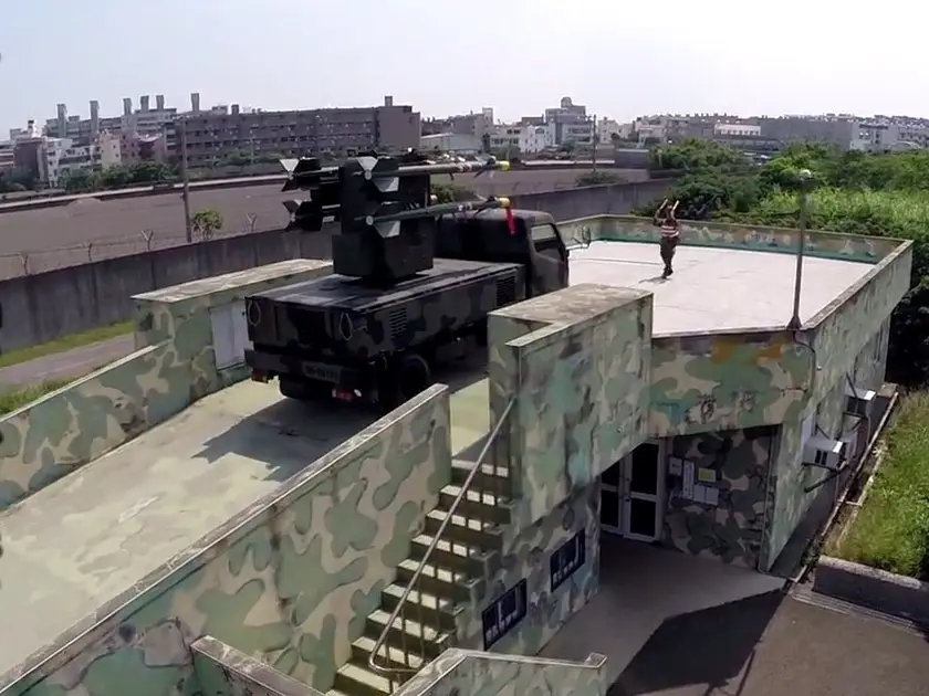 A military truck with a missile launcher is parked on a rooftop next to a camouflaged building. A person in casual clothes…