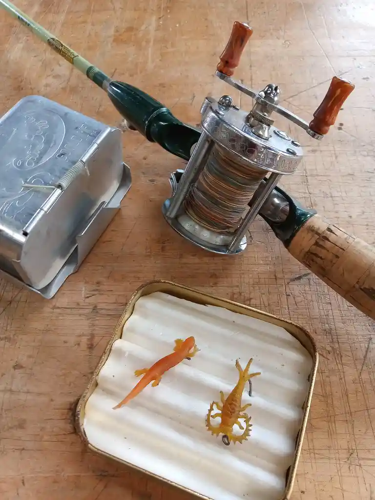 A vintage fishing rod with a silver reel sits on a wooden table next to a metal tin and a small open tin containing two…