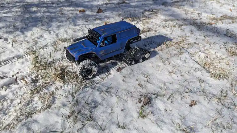 A blue remote-controlled toy truck with one wheel and one tracked rear end sits in a patch of snow and grass. The overlaid…