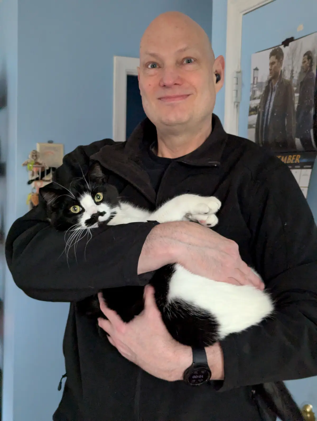 A bald man in a black jacket holds a black-and-white cat with wide, startled eyes. The man has a subtle, knowing smile and…