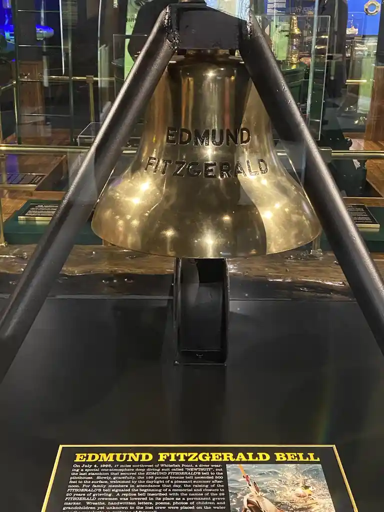 A large brass ship's bell inscribed with 'EDMUND FITZGERALD' is displayed in a museum case, suspended by black metal…