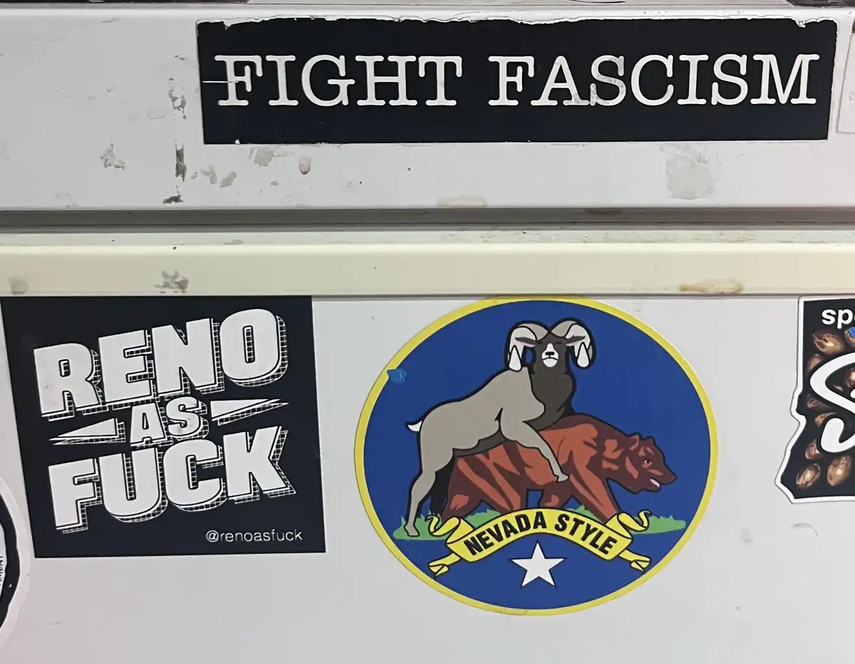 A close-up of a white surface covered in stickers, including a black sticker reading 'FIGHT FASCISM' at the top, a black…