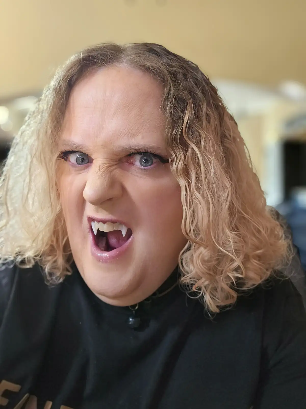 A person with curly blonde hair and a black shirt is making an exaggerated angry face with bared teeth that look like…