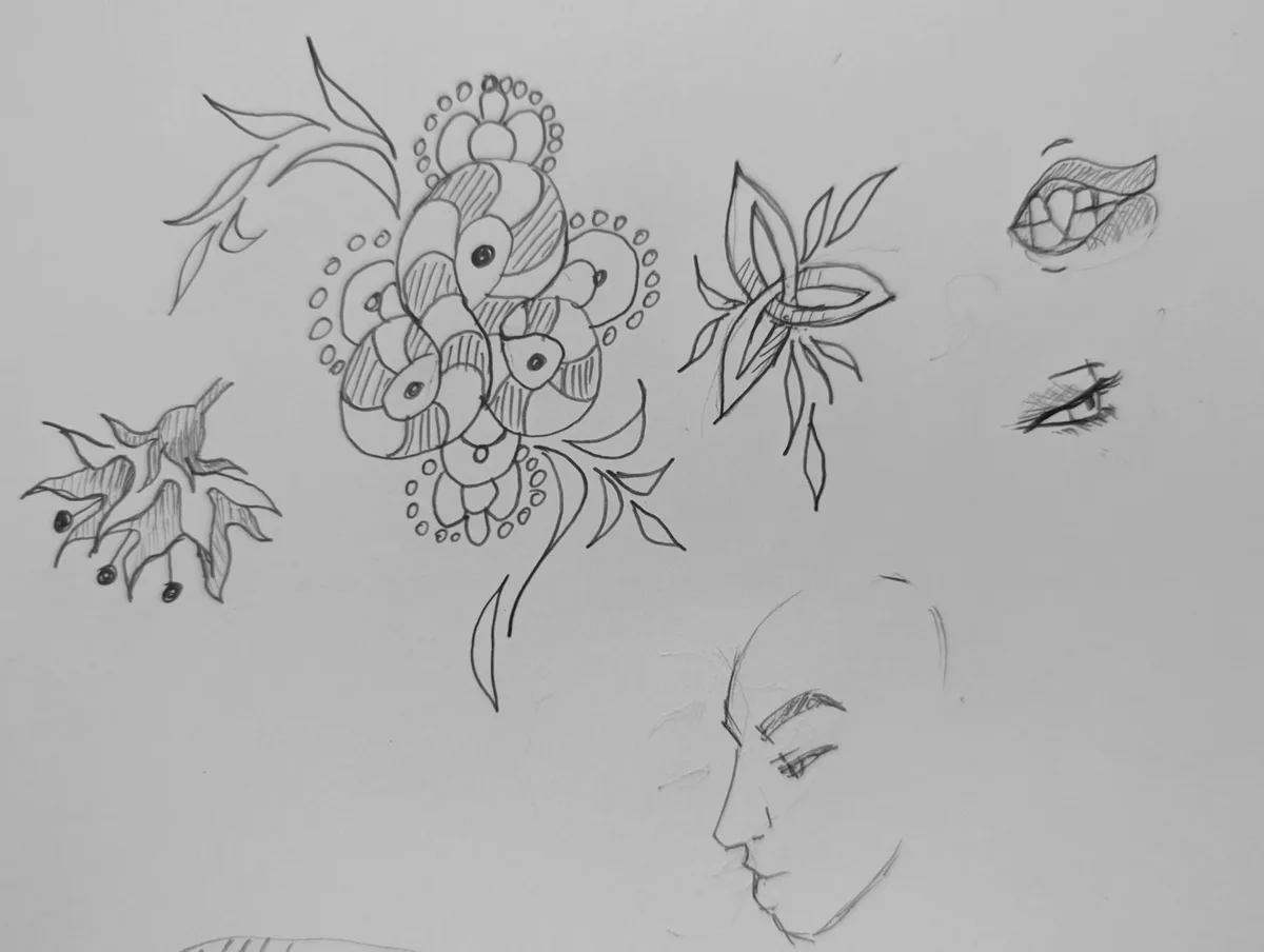 A pencil sketch on white paper shows various artistic doodles including a large central floral design, smaller flowers…