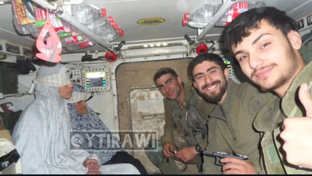 Inside a military vehicle, four men in olive green uniforms pose for a selfie. One man in the foreground gives a thumbs-up…