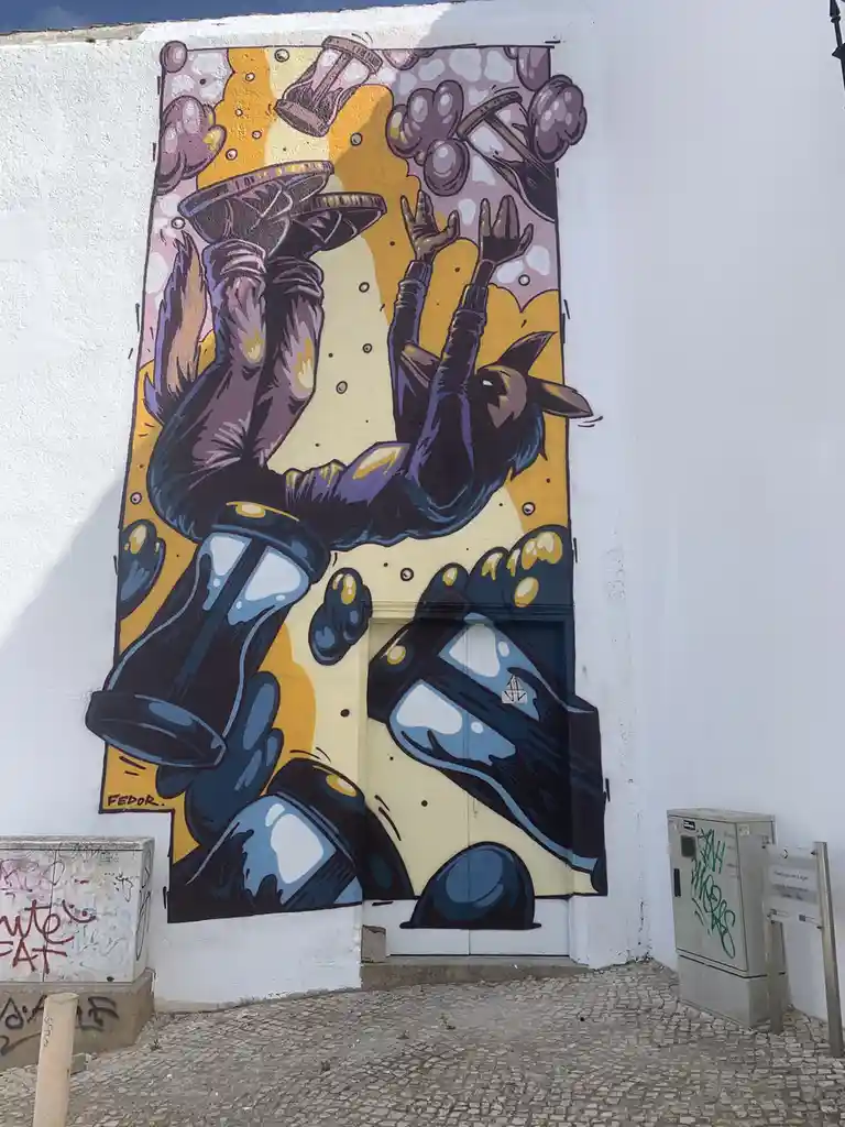 A vibrant street mural by artist FEDOR depicts a stylized, upside-down figure in a purple and blue outfit with a dog-like…