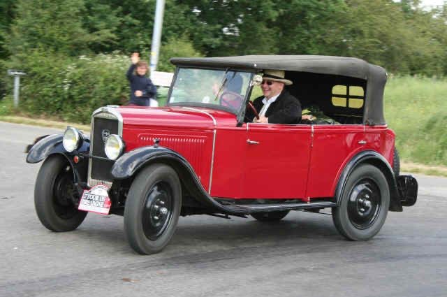 A man in a hat and sunglasses drives a red vintage convertible car with a black soft top. The car has a license plate that…