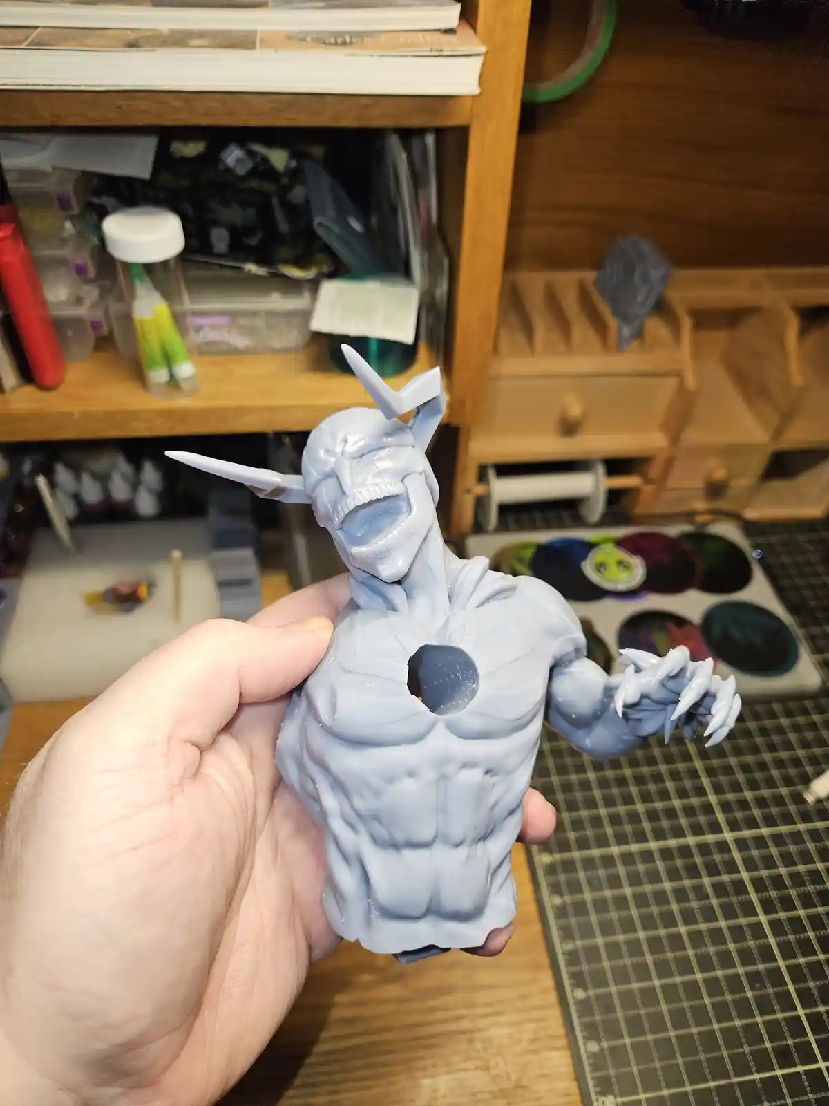 A person holds a grey-painted, unpainted action figure of a muscular, demonic-looking character with horns and claws…