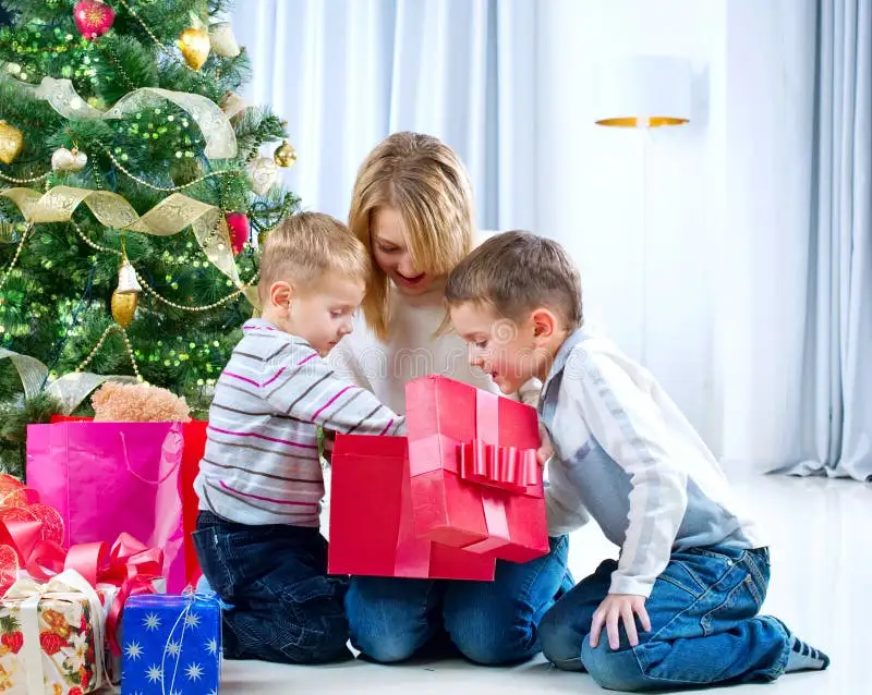 A woman and two young boys are kneeling on a white floor in front of a decorated christmas tree. They are gathered around a…