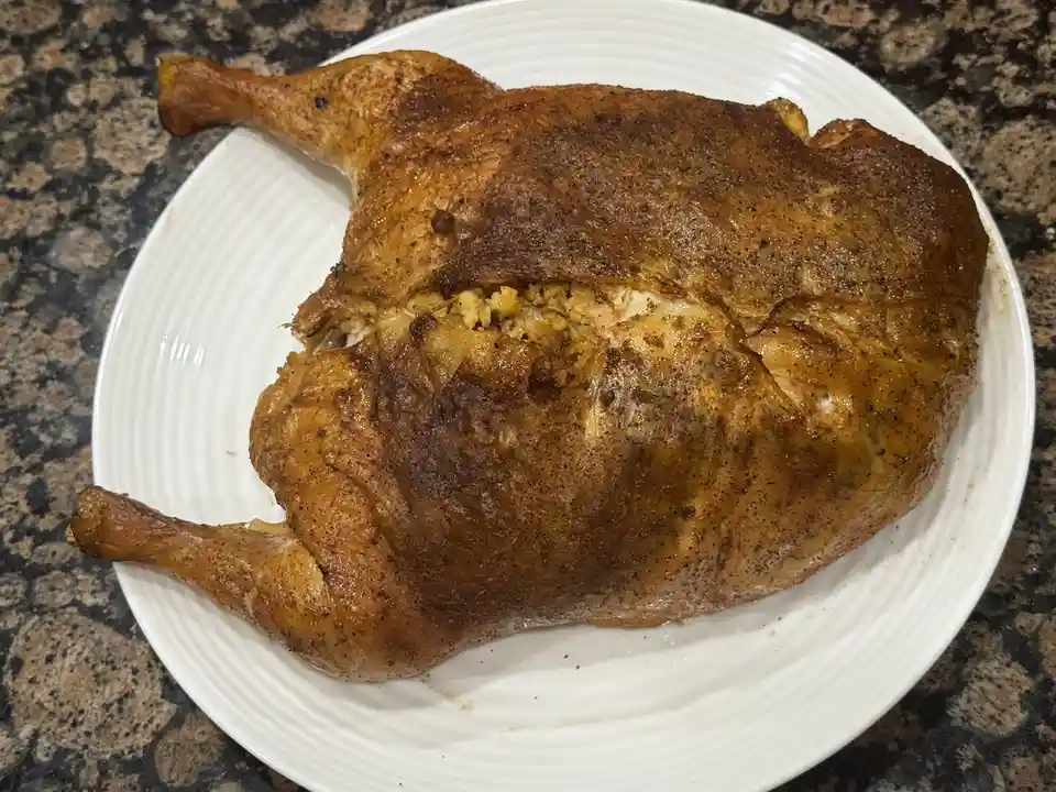 A whole roasted duck with golden-brown, seasoned skin sits on a white plate with ridged edges, placed on a dark speckled…