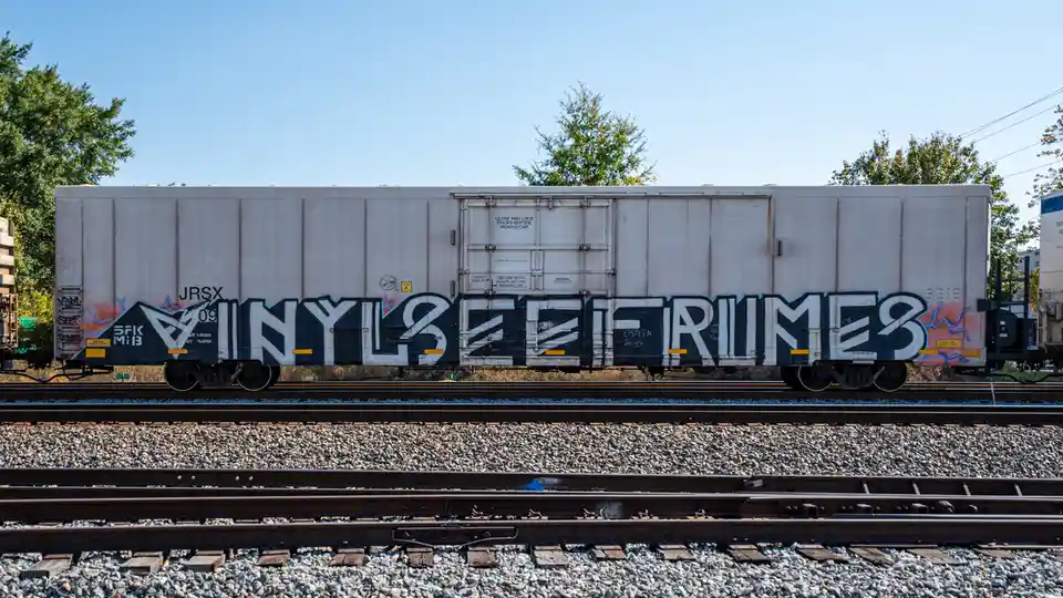 A white freight train car sits on tracks under a clear blue sky, covered in large black-and-white graffiti that reads…