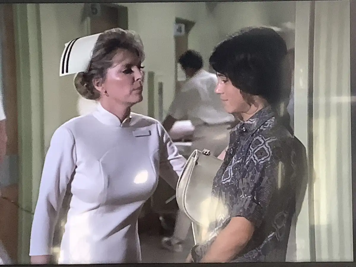 a still from a 1970s TV show shows a nurse in a white uniform and cap standing beside a woman in a patterned blouse holding…