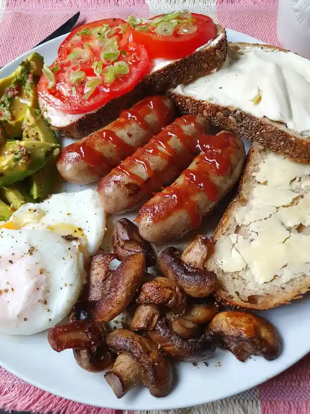 A plate of breakfast food including sausages with ketchup, fried mushrooms, poached eggs, avocado, and buttered toast with…