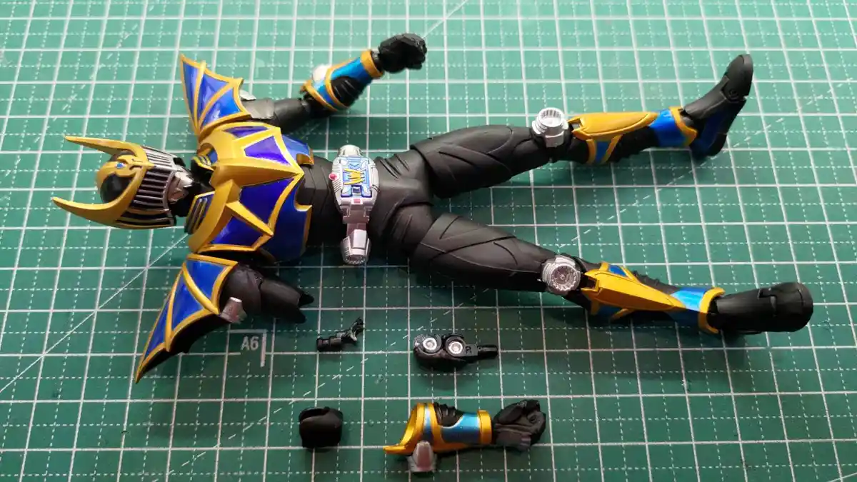 A disassembled Kamen Rider action figure lies on a green cutting mat with its limbs and accessories scattered around it. The…