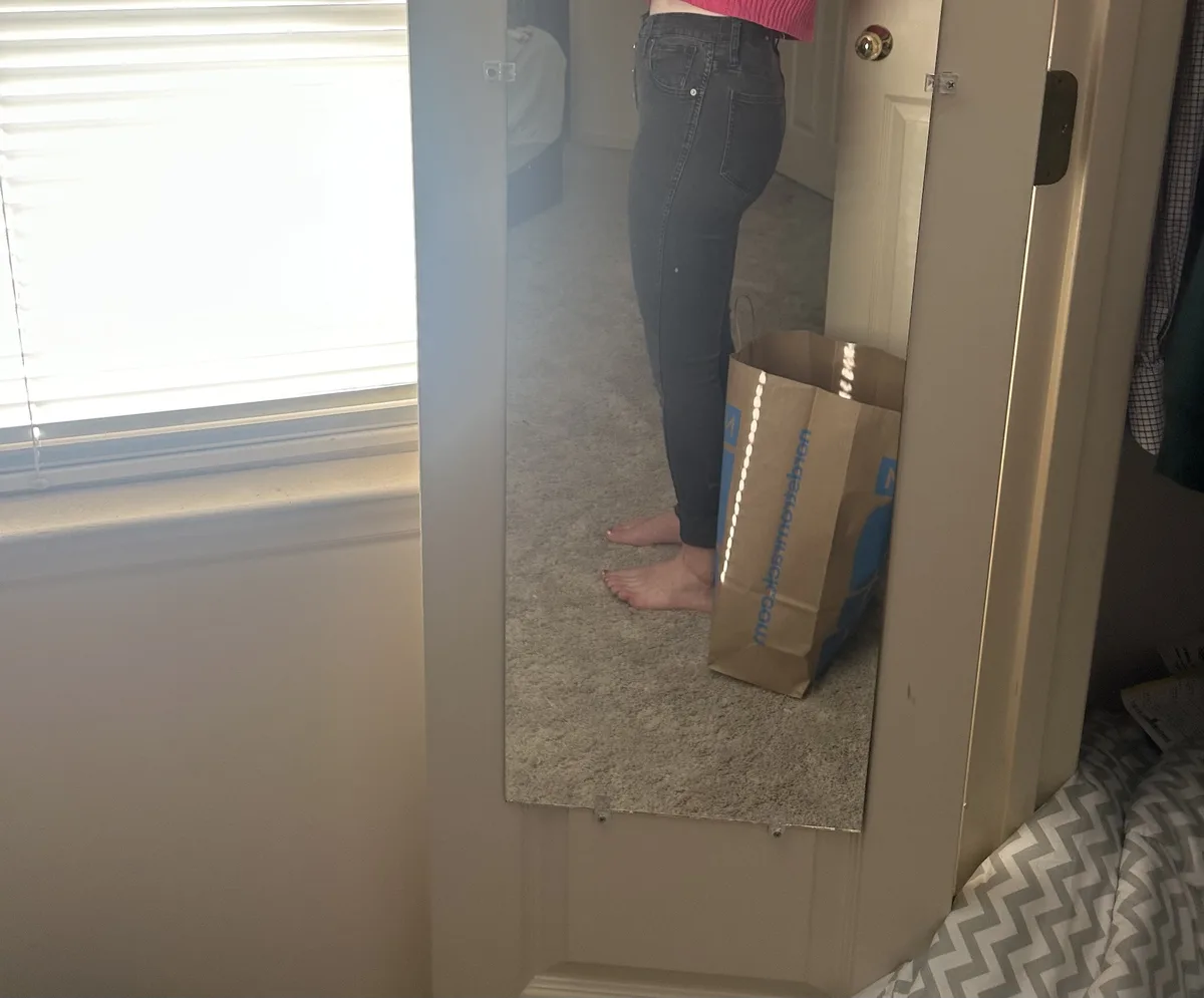A mirror selfie shows a person wearing dark jeans and a pink top, standing barefoot next to a Nordstrom Rack shopping bag…