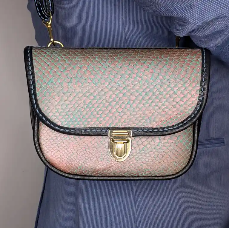 A close-up photo shows a person wearing a small, saddle-style handbag with a shimmering, iridescent snakeskin pattern that…