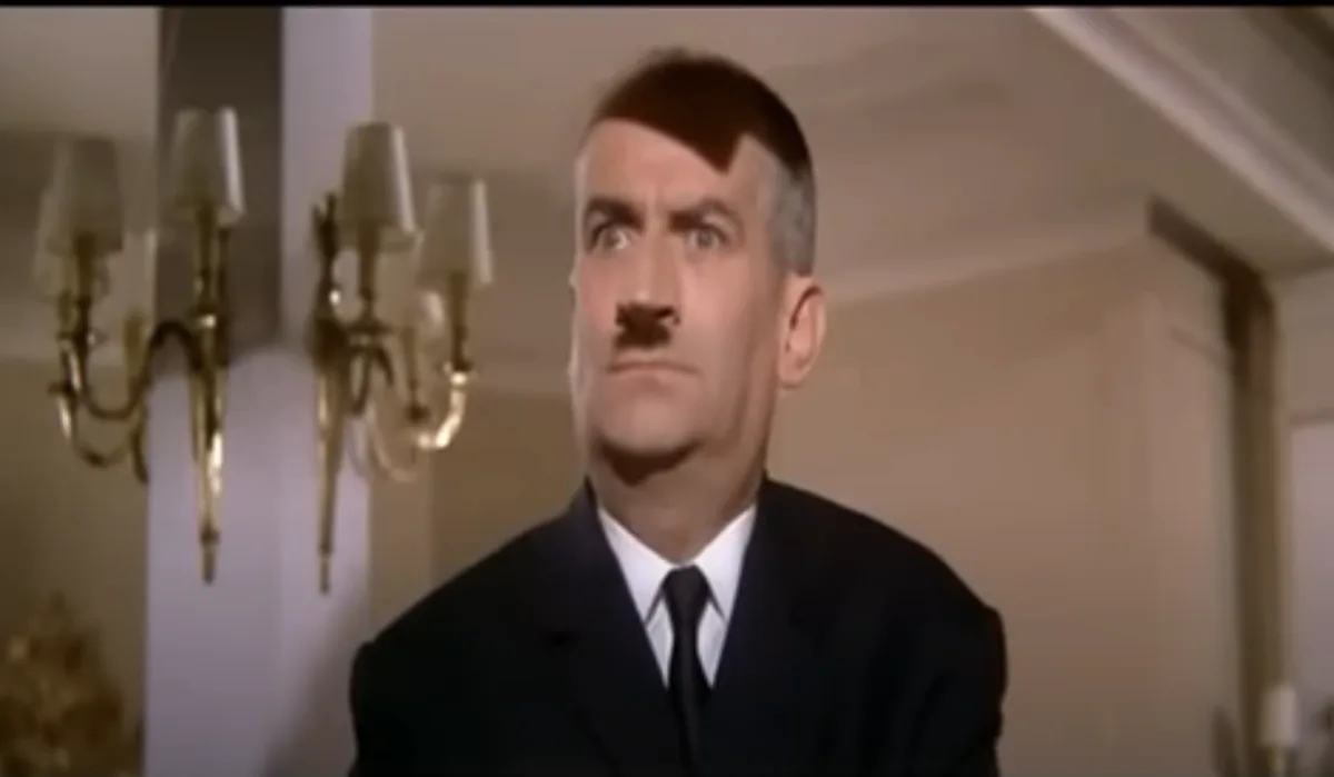 a man in a dark suit and tie with a mustache and a distinctive haircut looks upward with a shocked expression. Behind him…