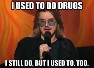 A man with long hair and sunglasses holds a microphone against a fiery orange background. The text overlaid in white capital…