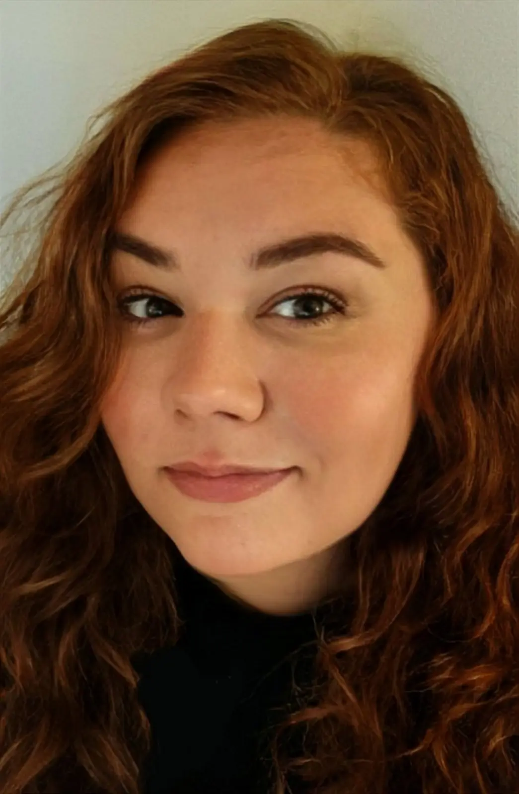 A close-up portrait of a young woman with long, wavy auburn hair and a subtle smile, wearing a black turtleneck against a…