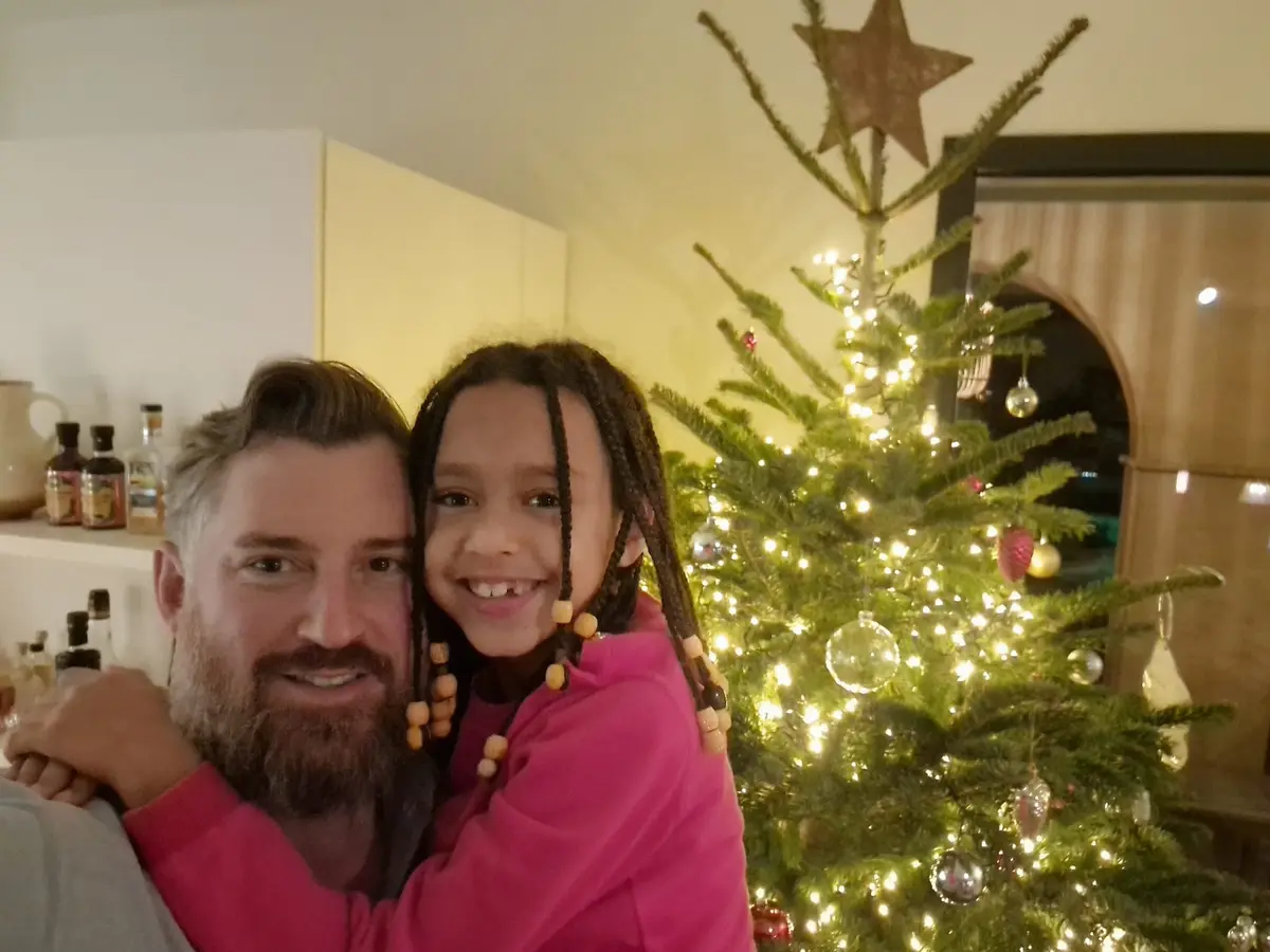A man with a beard and a young girl with braided hair, both wearing pink, are smiling and hugging in front of a lit…