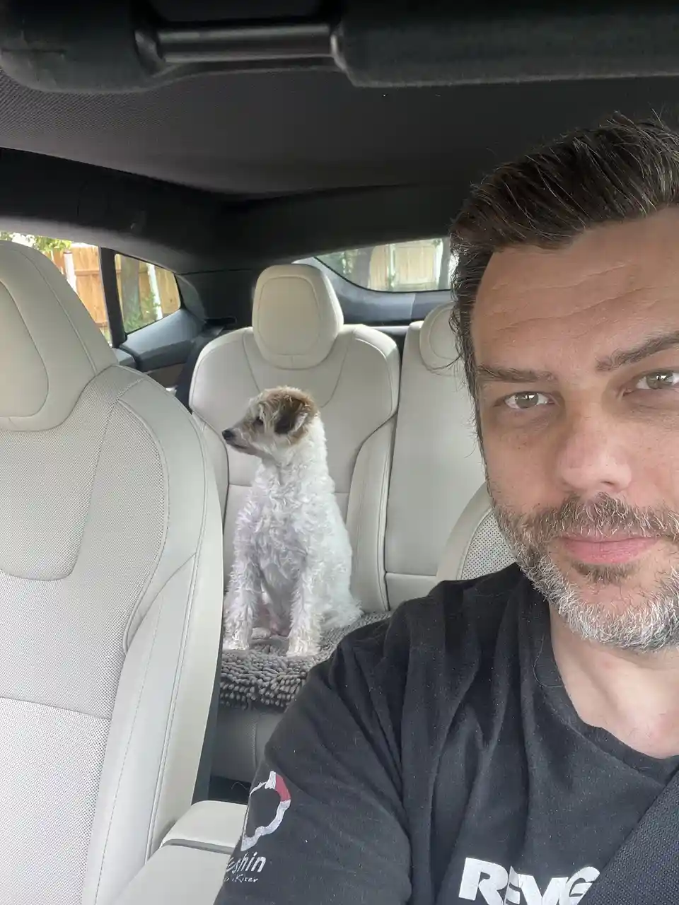 A man takes a selfie in the driver's seat of a car with light-colored leather seats, looking directly at the camera with a…