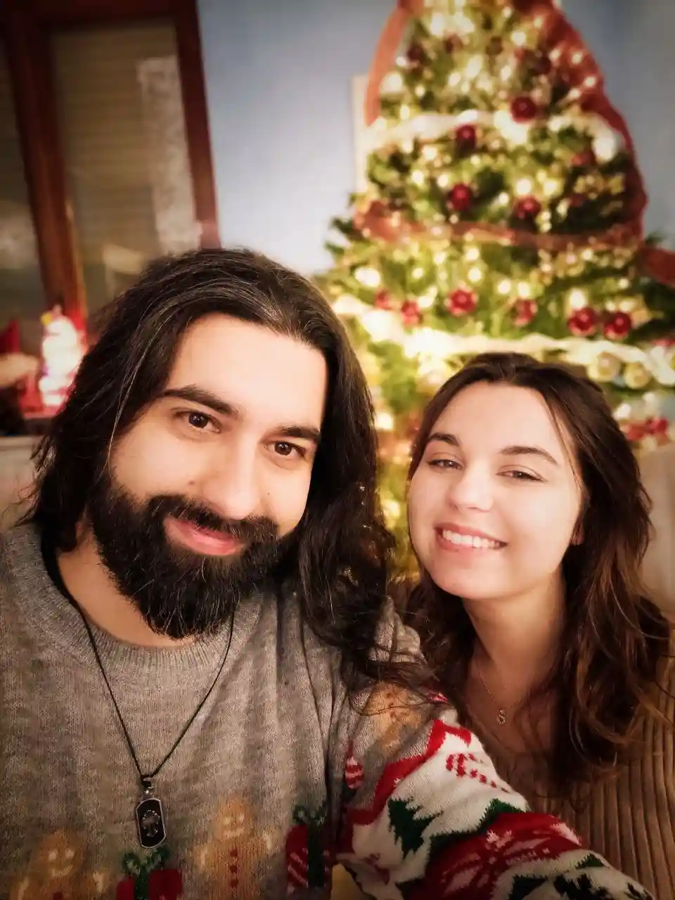 A man and woman pose for a selfie in front of a decorated Christmas tree. The man has long dark hair and a beard, wearing a…