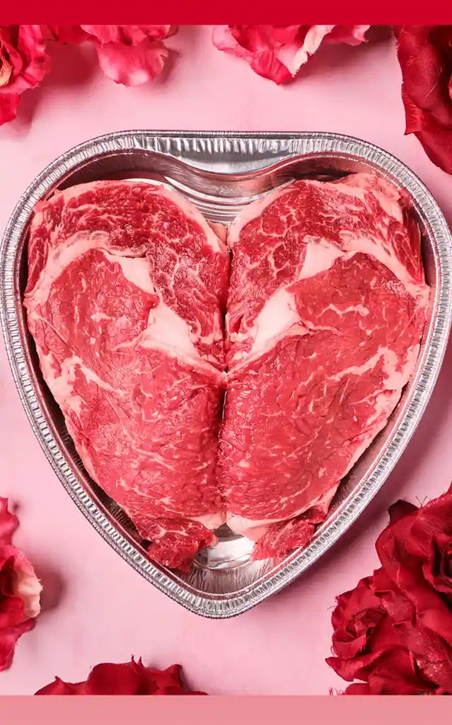 Two raw beef steaks are arranged in a heart shape inside a heart-shaped aluminum tray, surrounded by red roses on a pink…