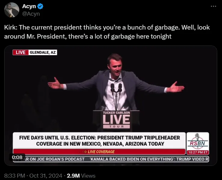A man in a suit stands at a podium labeled 'Tucker Carlson Live Tour' with arms spread wide, seemingly addressing an…