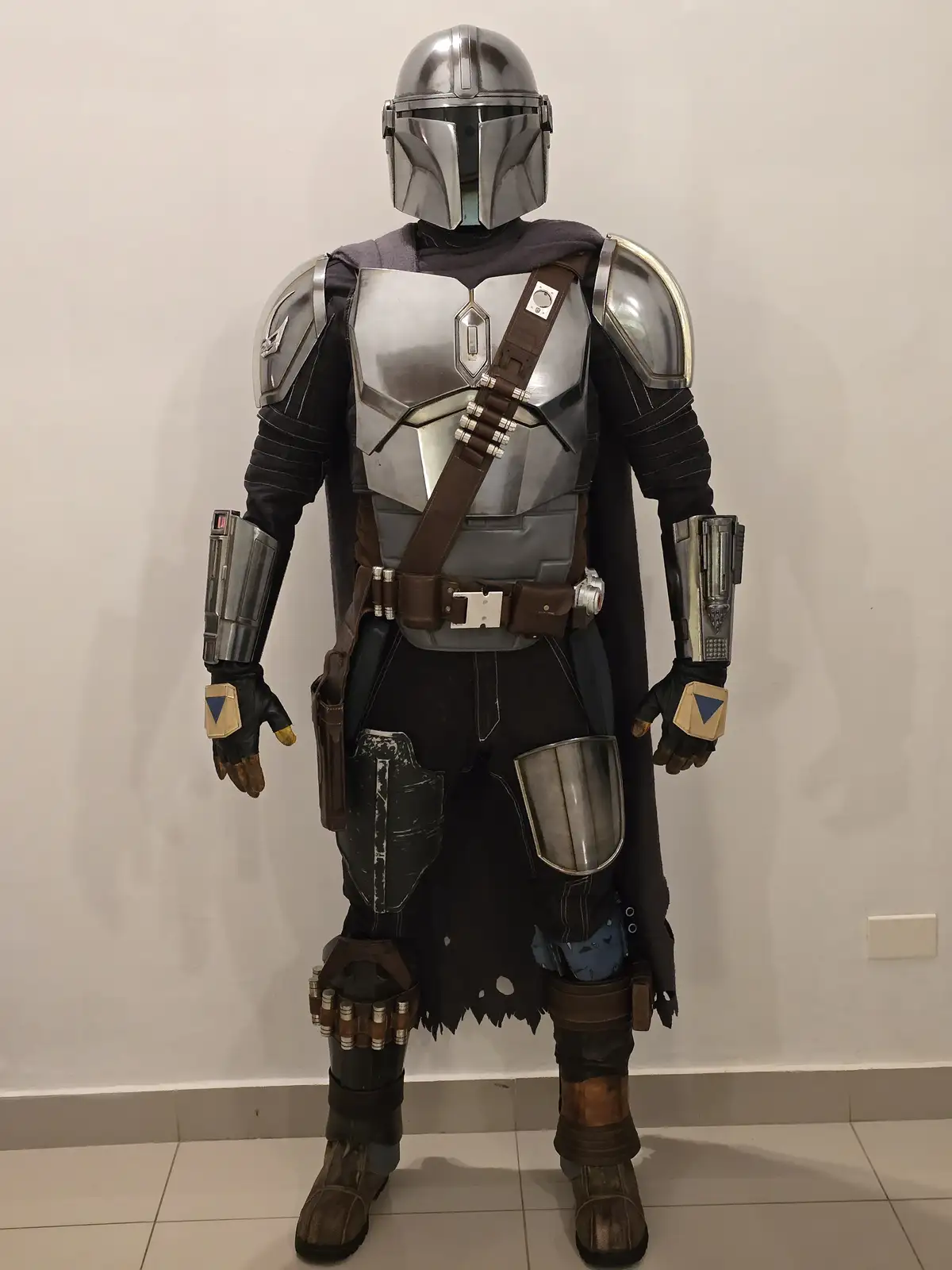 A person in a full-body Mandalorian armor costume, complete with helmet, chest plate, shoulder pads, gauntlets, and a dark…