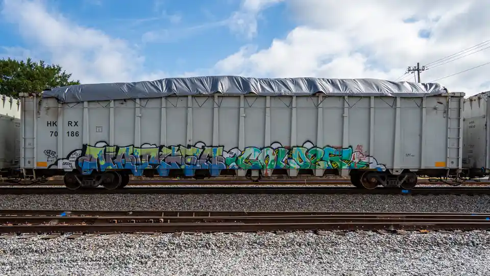 A side view of a white freight train car covered with a gray tarp, sitting on railroad tracks under a partly cloudy sky. The…