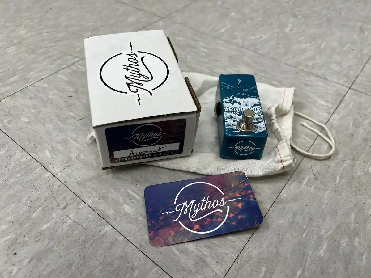 A blue Mythos Argonaut guitar effects pedal sits next to its white cardboard box, a white drawstring bag, and a branded…