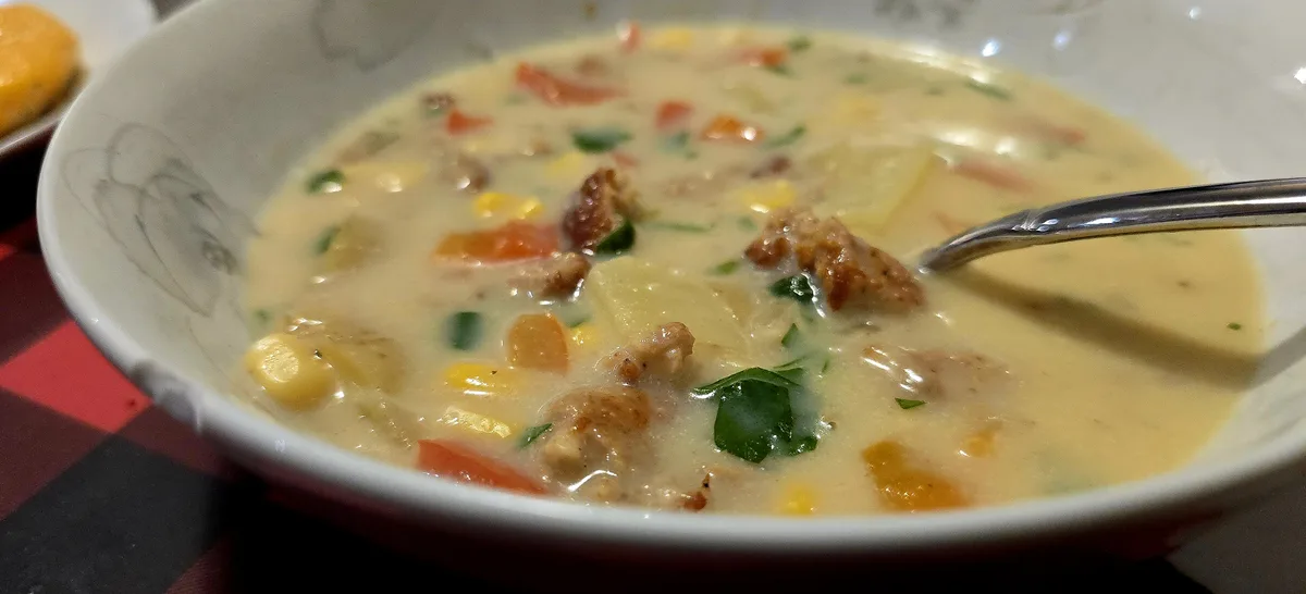 A close-up photo of a creamy, chunky soup in a white bowl with a spoon, containing visible pieces of corn, potatoes…