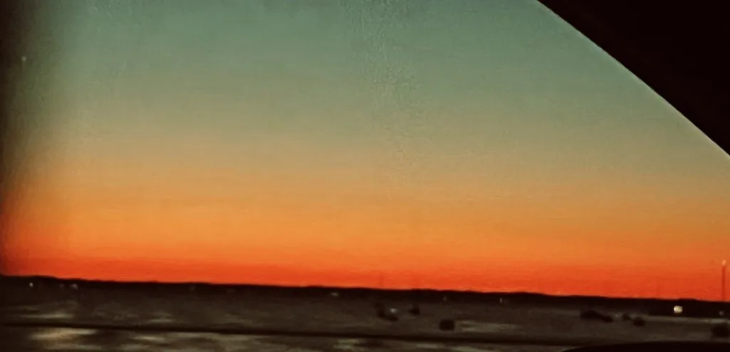 A grainy, vintage-style photo shows a sunset over water, viewed from inside a vehicle with a visible window frame. The sky…
