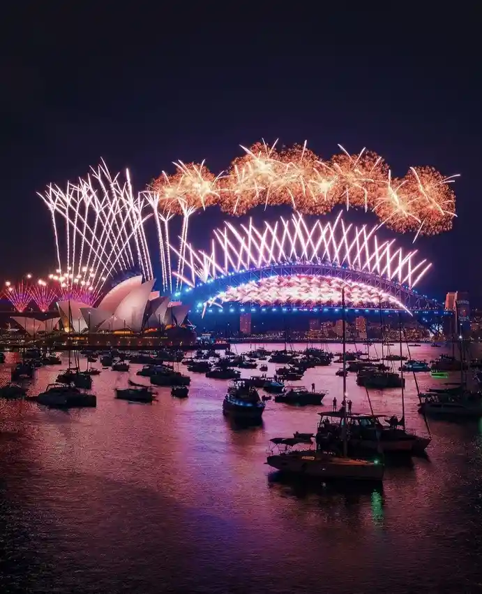 A nighttime photograph of Sydney Harbour with the Sydney Opera House and Harbour Bridge illuminated, fireworks exploding…
