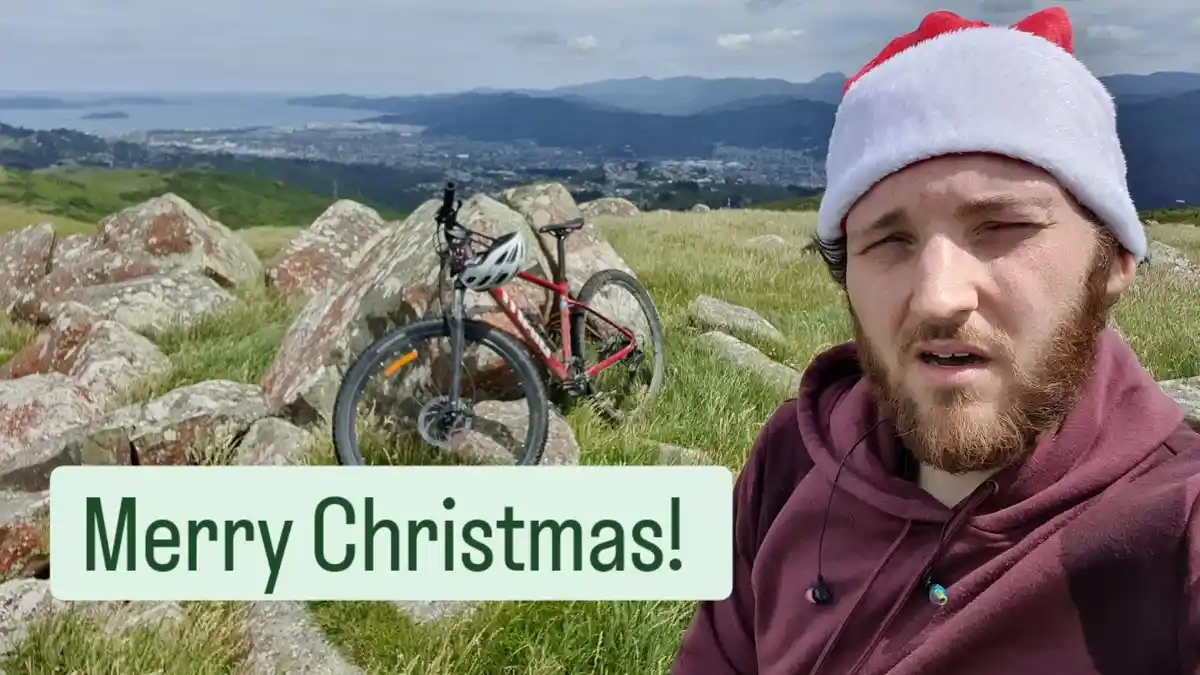 A man with a beard wearing a red and white Santa hat and a maroon hoodie stands on a grassy hillside with large rocks…