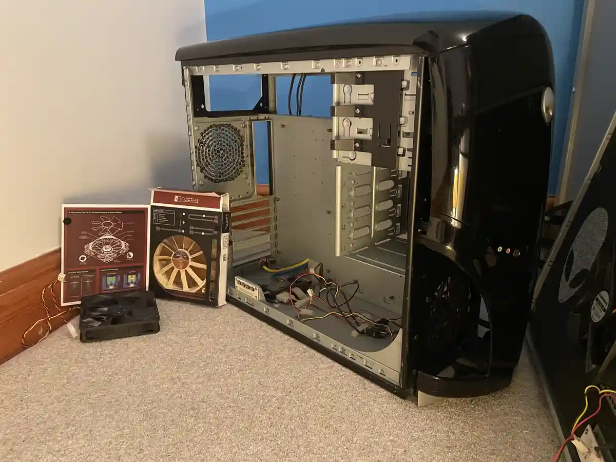 An open black computer case sits on a carpeted floor, surrounded by uninstalled components including a Noctua fan box, a fan…
