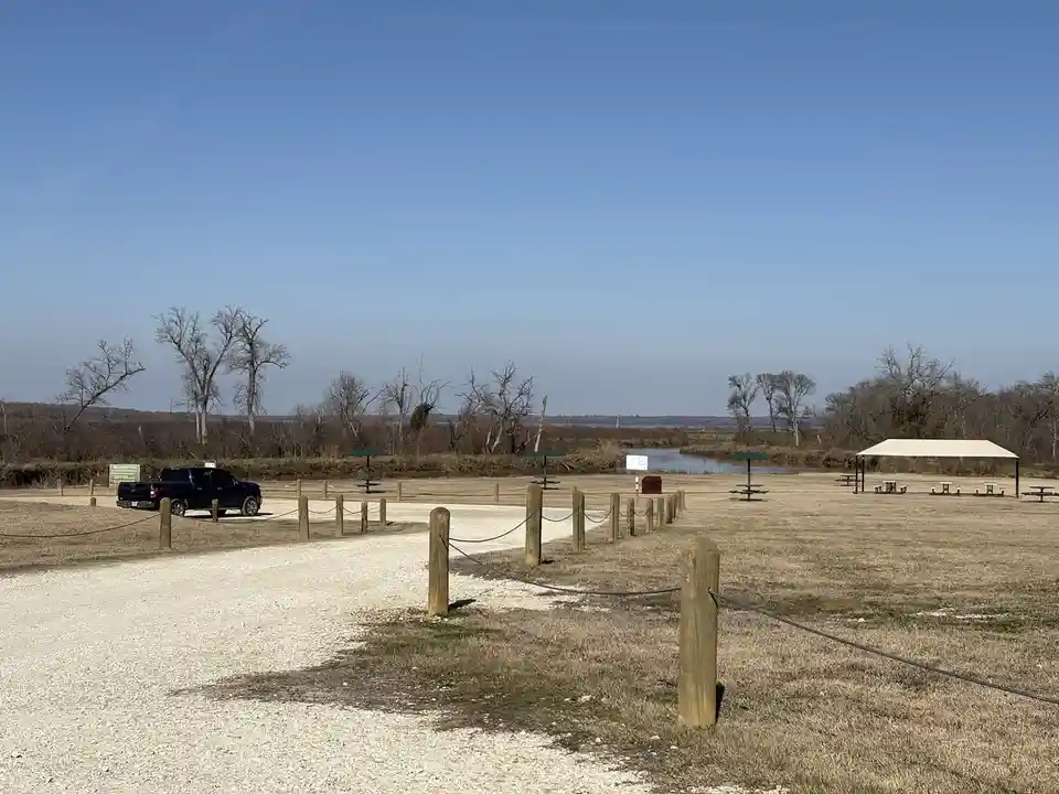 A wide shot of a quiet park with dry grass, a gravel path, picnic tables, and a single black pickup truck parked near a…