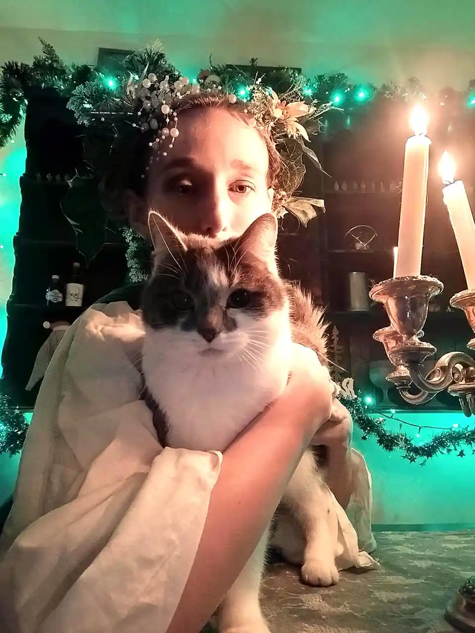 A person wearing a floral crown and draped in a light-colored robe holds a fluffy white and gray cat in front of them. The…