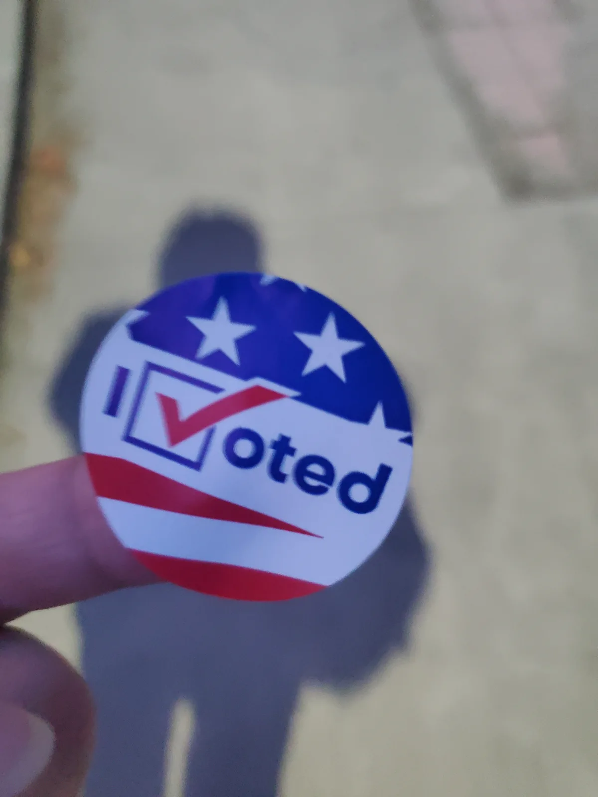 A close-up photo shows a person's thumb holding a circular 'I Voted' sticker, which features stars and stripes in red…