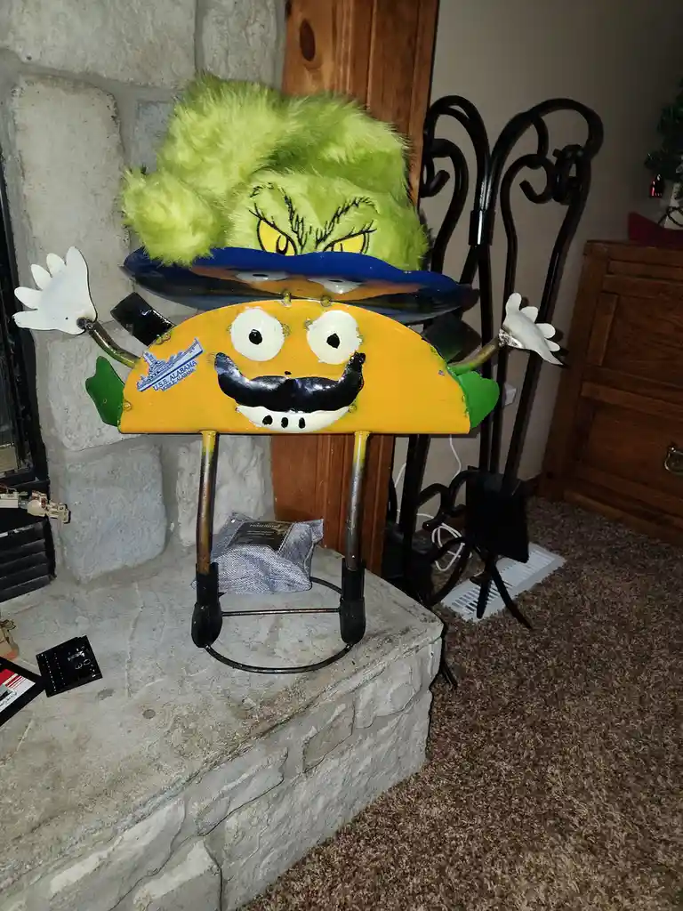 A handmade, whimsical sculpture sits on a stone fireplace mantel, crafted to look like a cheerful yellow taco with a black…