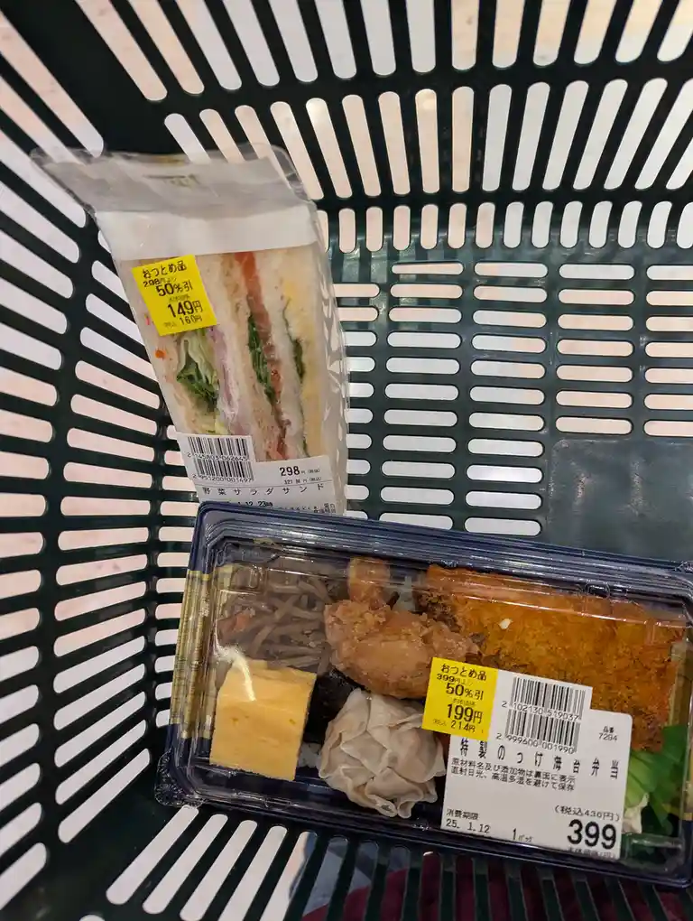 Two discounted Japanese convenience store meals sit in a black plastic shopping basket. The top item is a sandwich in a…